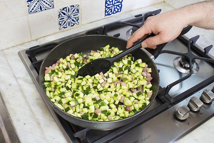 Aggiungete adesso le zucchine tagliate a cubetti piccoli, salatele e pepatele a piacere e saltatele un paio di minuti con gli altri ingredienti per farle ben insaporire. Versate un bicchiere d'acqua nella padella, mettete il coperchio e  lasciate cuocere le zucchine per una decina di minuti.