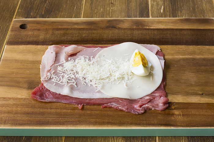 Stendete una fettina di carne sul tagliere e farcitela con una fetta di prosciutto, la provola, uno spicchio di uovo sodo e una spolverata di formaggio grattugiato. 