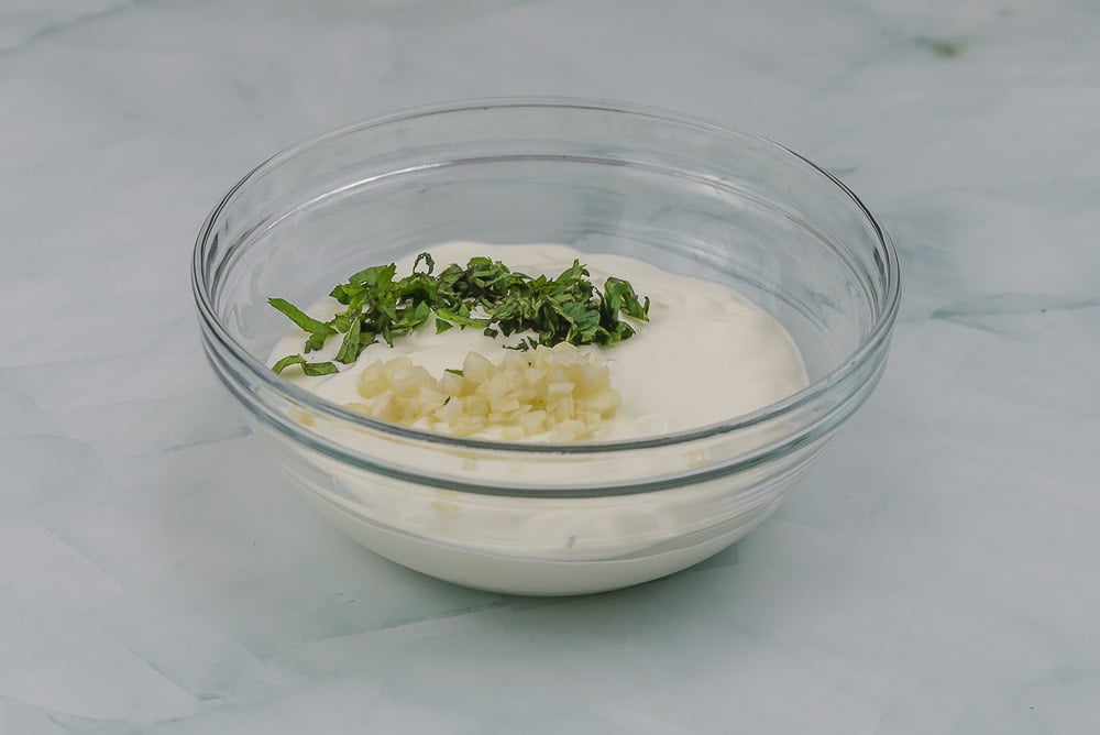 In una ciotola mettiamo lo yogurt greco, l’aglio tritato e la menta sminuzzata e mescoliamo.